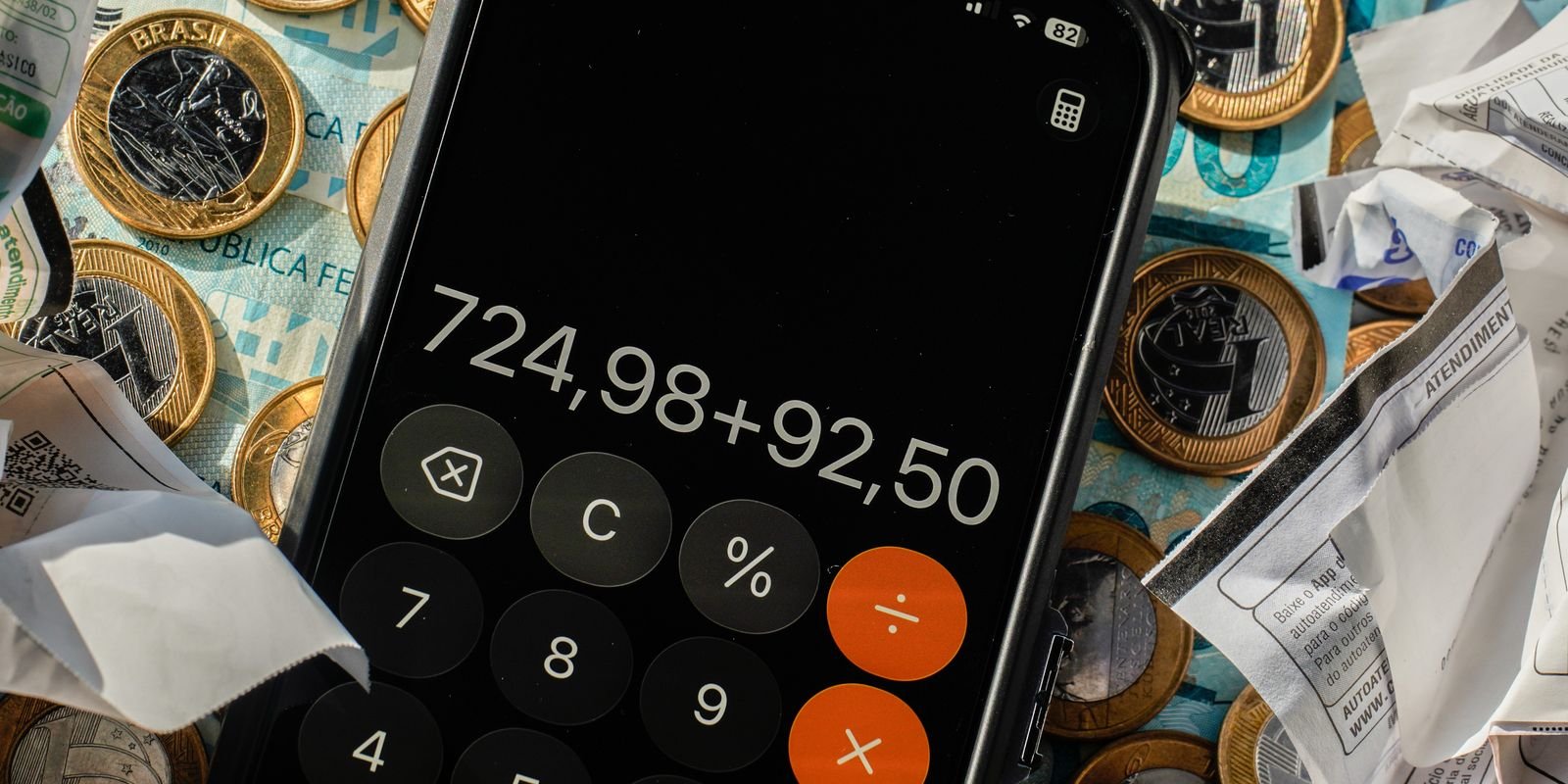 Calculadora de celular sobre moedas e cédulas de real e notas fiscais amassadas, em Brasília